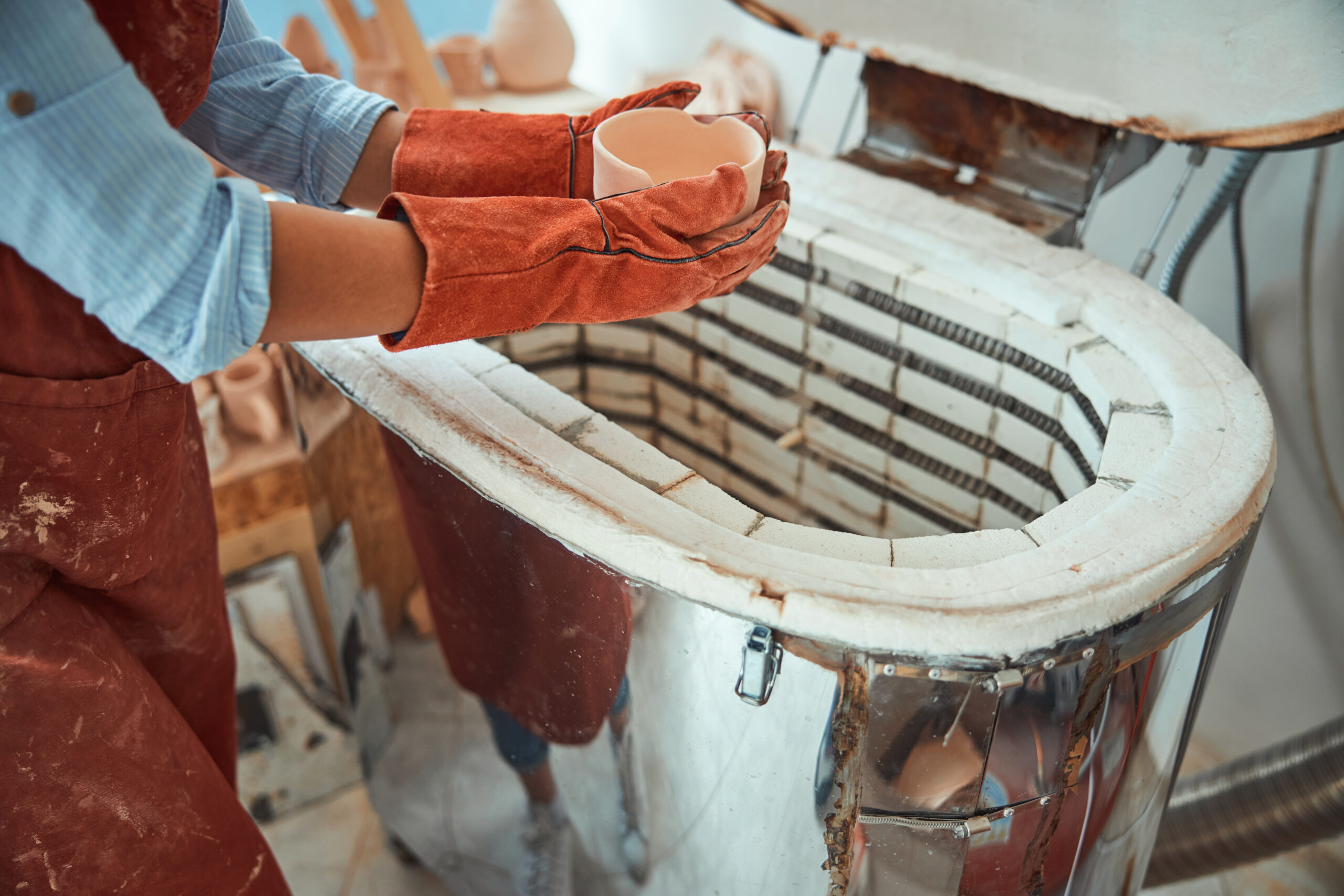Female potter in gloves using electric pottery kiln in workshop