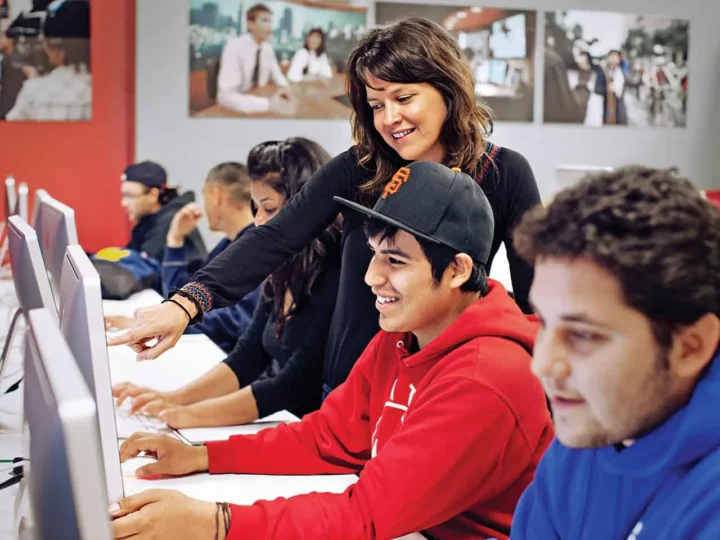 Students work on computers while a teacher points at one screen, instructing them. Classroom setting with educational posters on the walls.