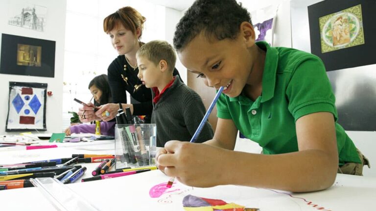 Children draw at a table with art supplies while a teacher observes. The classroom features art on the walls, suggesting a creative, educational environment.