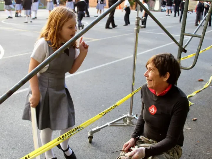 A woman kneels, smiling, interacting with a girl standing nearby. They are near metal scaffolding and yellow caution tape on a school playground, with other children in the background.