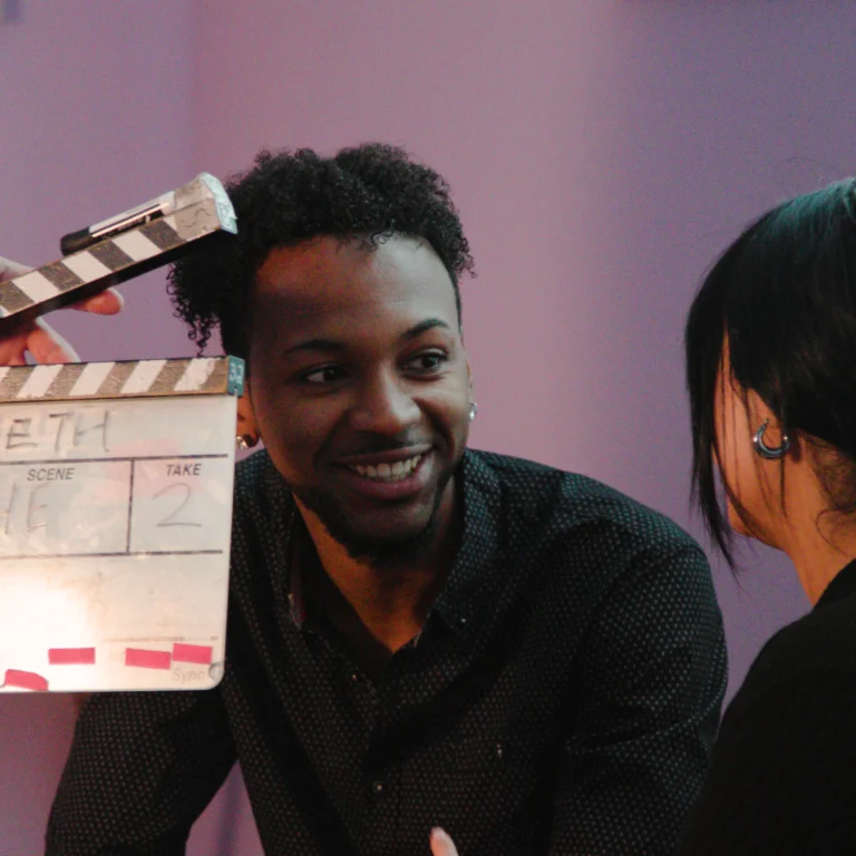 A smiling man sits across from a woman on a film set, engaged in conversation. A clapperboard is held in front of him, indicating the start of a scene. The background is lit with soft purple lighting, creating a relaxed atmosphere.