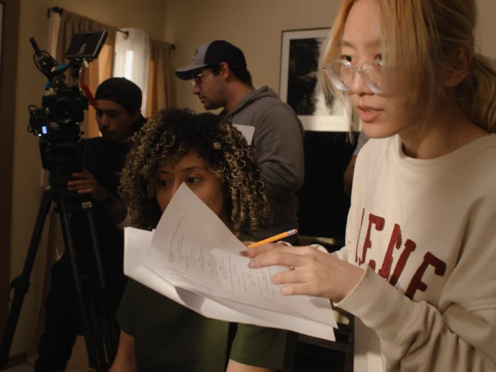 Film crew members work together indoors, reviewing a script while preparing to shoot a scene with a camera set up in the background.