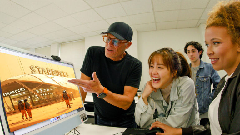 A group of five people looking at a computer screen displaying a design for Starbucks, with one person explaining while others watch and smile.