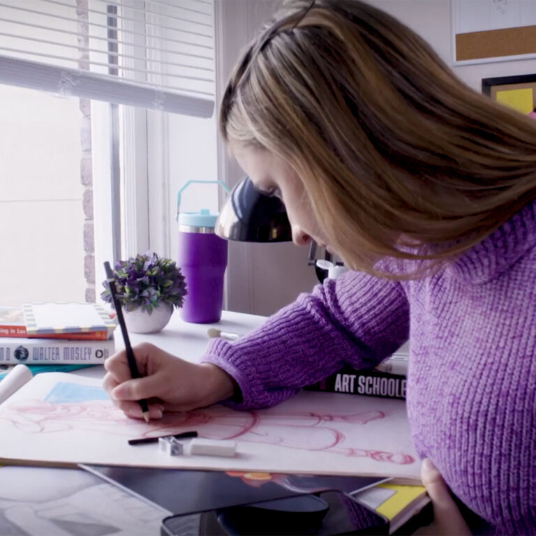 A girl in a purple sweater is drawing with a black pen on a large sheet of paper at a cluttered desk.