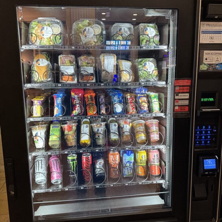 A vending machine stocked with salads, sandwiches, snacks, and canned beverages, including Monster and Coca-Cola options.