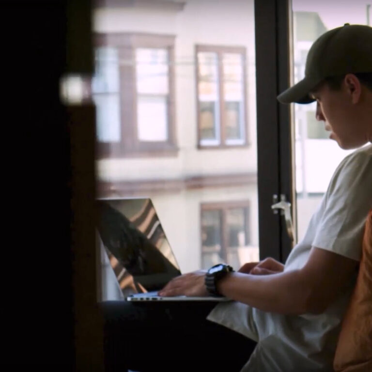 A young man wearing a cap and white t-shirt is sitting on a couch, working on a laptop near a window with natural light.
