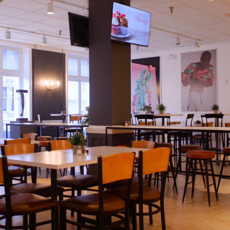 Empty cafe with tables, chairs, potted plants, large posters, and a TV display showing cupcakes and 