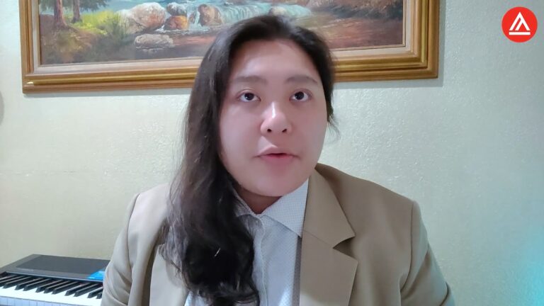 Man in beige blazer speaking, with a painting and electronic keyboard behind him.