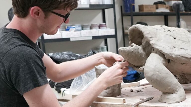 A person sculpts a large clay turtle on a wooden table, in an art studio with shelves containing various materials in the background.
